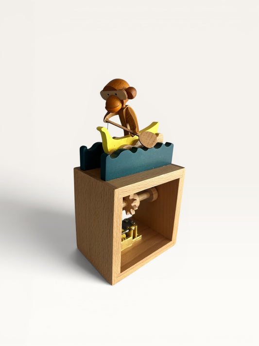 Juguete mecánico de madera Retrópica Tienda con figura de mono remando en canoa amarilla sobre olas azules.